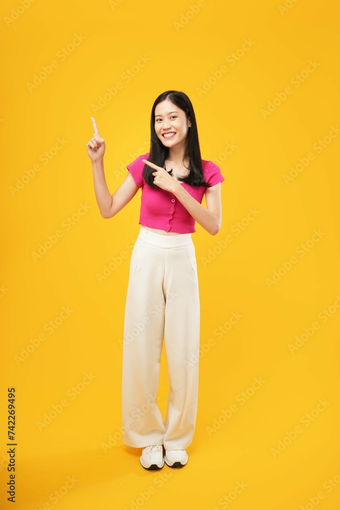 Full-length portrait of amazed excited young Asian woman 20s in a pink casual t-shirt pointing index fingers aside up on mock-up copy space isolated on Yellow color wall background studio Full-length portrait of amazed excited young Asian woman 20s in a pink casual t-shirt pointing index fingers aside up on mock-up copy space isolated on Yellow color wall background studio