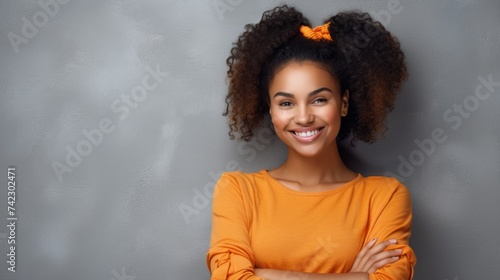 A young happy African American woman wearing an orange sweatshirt, smiling with her arms crossed on her chest on a gray background with a copy space. © liliyabatyrova