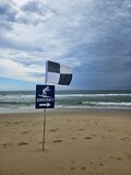 Cloudy Beach Day: black and white flag marks designated surfcraft area, waves gently crashing as footprints scatter across the sandy shore