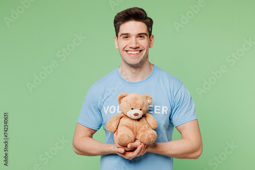 Young man wears blue t-shirt white title volunteer hold hug teddy bear plush toy look camera isolated on plain pastel light green background. Voluntary free work assistance help charity grace concept.