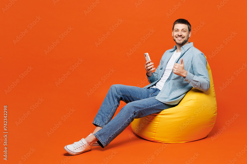 Full body young fun man he wears blue shirt white t-shirt casual clothes sit in bag chair use mobile cell phone show thum up isolated on plain red orange background studio portrait. Lifestyle concept. Full body young fun man he wears blue shirt white t-shirt casual clothes sit in bag chair use mobile cell phone show thum up isolated on plain red orange background studio portrait. Lifestyle concept.