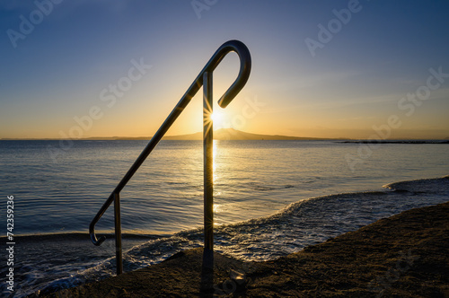 Handrail framing sunrise over the top of Rangitoto Island. Milford Beach. Auckland.