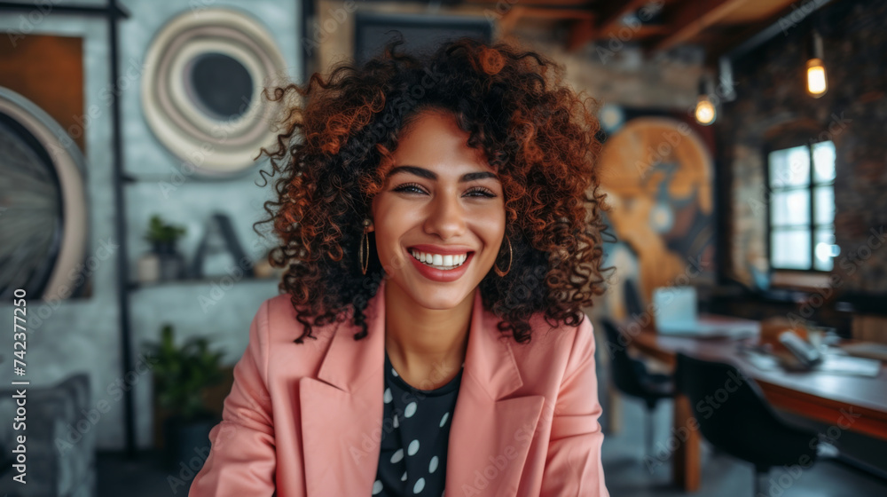 A vibrant portrait of a Gen Z businesswoman laughing heartily, her curly hair and bright pink blazer adding a pop of color and energy against the creative backdrop of a modern workspace