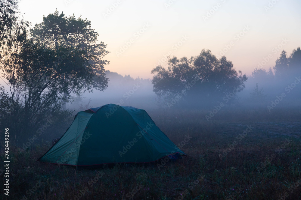 Green tourist tent by the river at sunrise, with morning autumn fog on the water. Outdoor tourist landscape.