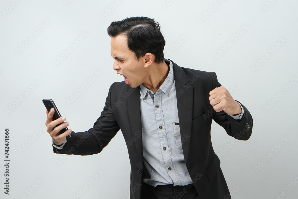 angry mad asian indonesian business man in suit holding smart phone on isolated background angry mad asian indonesian business man in suit holding smart phone on isolated background