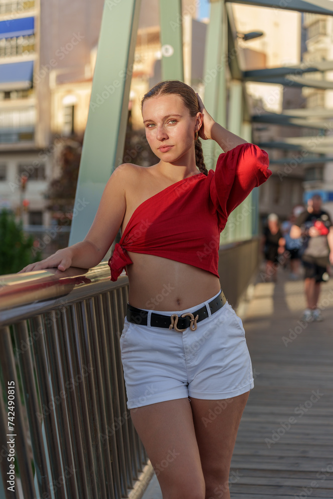 Sensual girl dressed in white shorts and a red one-shoulder top, posing on the railing of a bridge with the city and people out of focus in the background.
