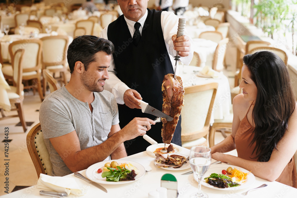 Happy couple, date and dining with kebab of waiter serving, meat or slices on romantic dinner at table. Young man or woman with chef, skewer or food for meal, eating or enjoying service at restaurant