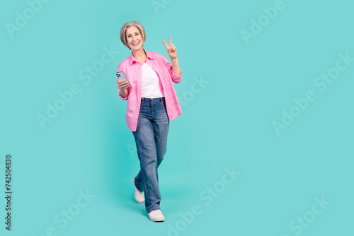 Full length photo of cheerful senior woman wear pink shirt holding smartphone go empty space show v-sign isolated on teal color background