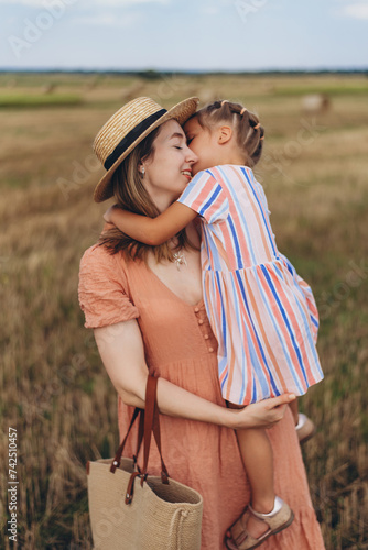 A young mother in a straw hat and with a wicker bag holds a small daughter in her arms. Both stand in a field of harvested wheat. Against the background of a bale of straw