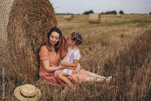 Mother and daughter sitting near a bale of straw on a harvested wheat field. against the background of other bales. Daughter touches mom by her hair