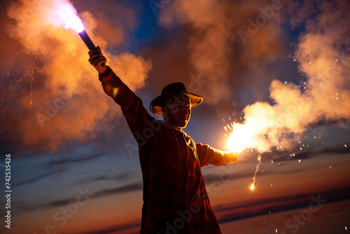 Attractive man is standing with open arms and holding bright signal fire against sunset sky photo