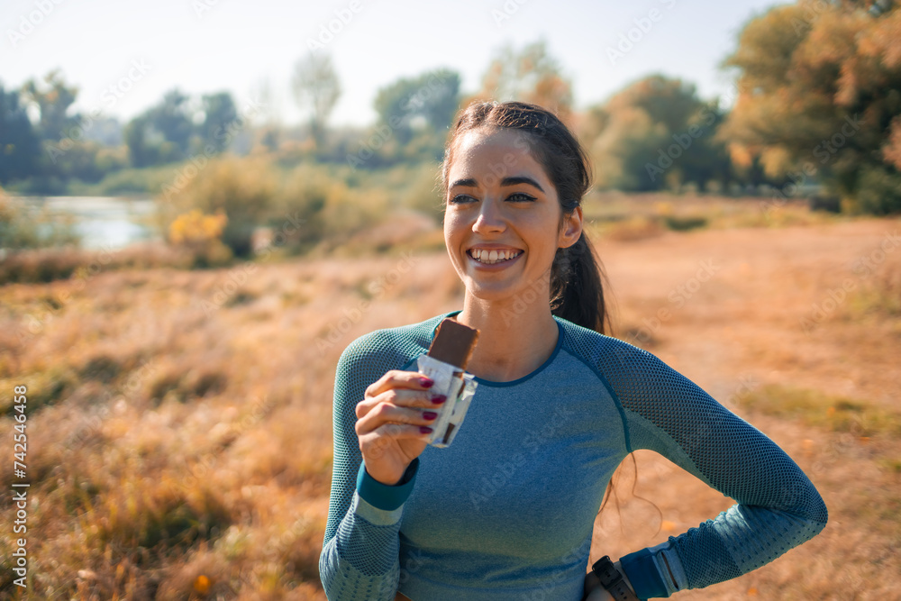 A satisfied young female athlete taking a break from her routine outdoor jog and having a protein bar as a snack