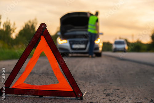 Unrecognizable sad driver in reflective vest. Male driver standing near broken car with open up hood. Red triangle to warn other road users of car breakdown or engine failure stop at countryside