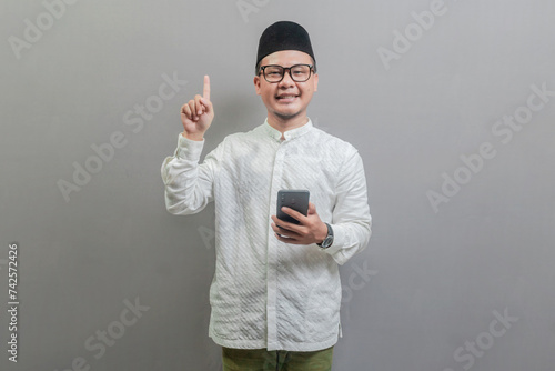 Portrait of an Asian Muslim man wearing a koko shirt and peci with shades of the fasting month, smiling pointing finger to up free space and holding his smartphone, isolated on a white background