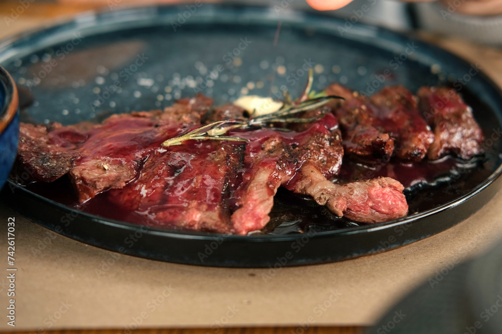 Man Cutting Rib Eye Steak, Pouring it Red Sauce on a Black Plate. Person Having a Delicious Lunch. Grilled Sirloin Beef Steak with Rosemary and Garlic on a Wooden Table. BBQ Meat Delicacy Bio Food.