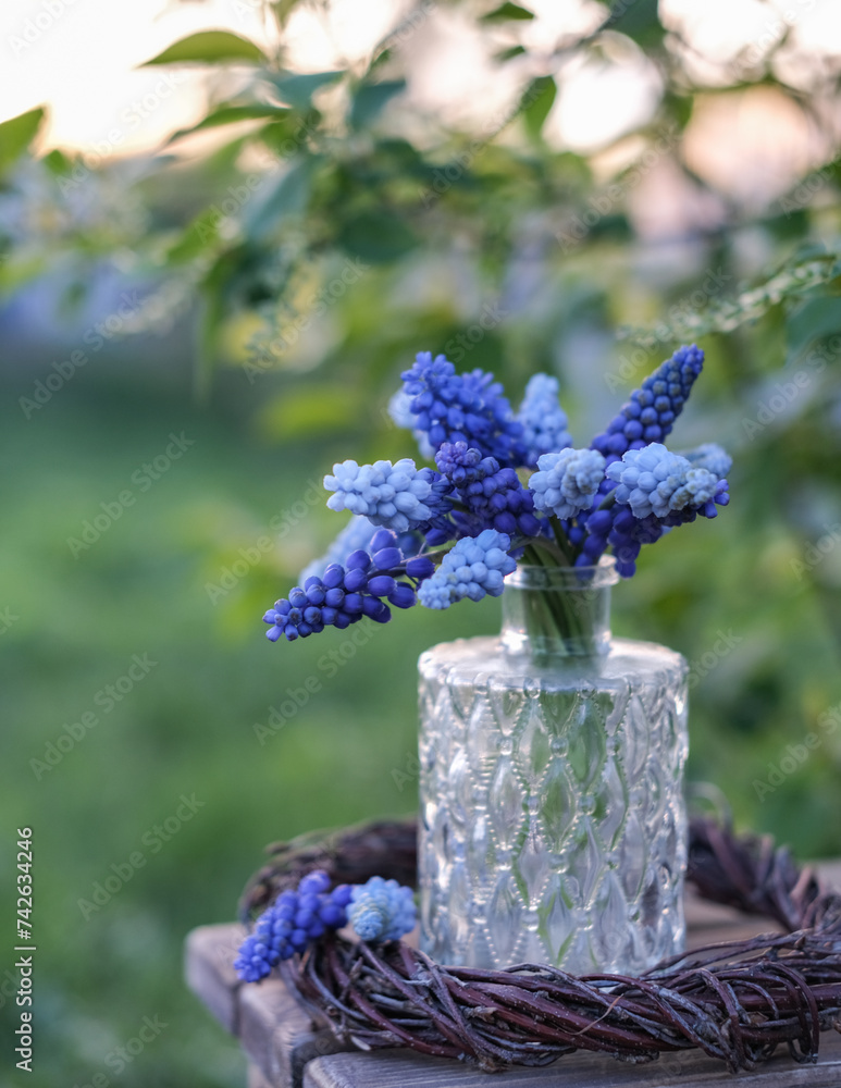 Blue grape hyacinths in a clear patterned vase on a woven wreath, with a soft-focus garden background
