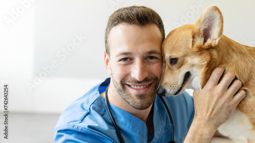 Pet care concept. Friendly male veterinarian playing with cute pembroke welsh corgi dog during checkup at clinic, man looking at camera and smiling, copy space