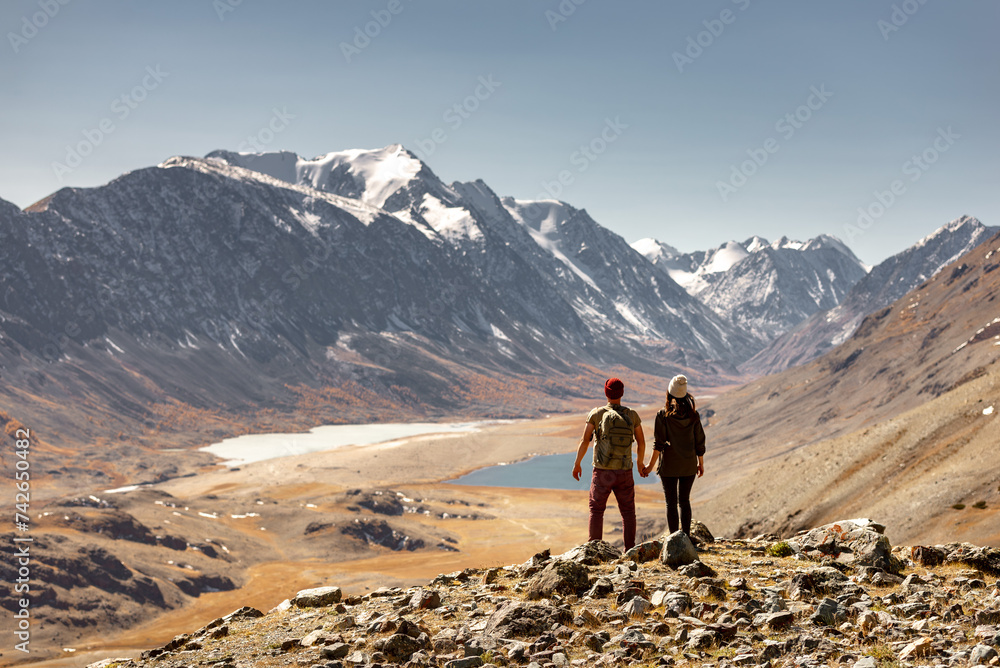 Couple of tourists or hikers are standing at mountain top and enjoys nice view at valley with two lakes and small glaciers