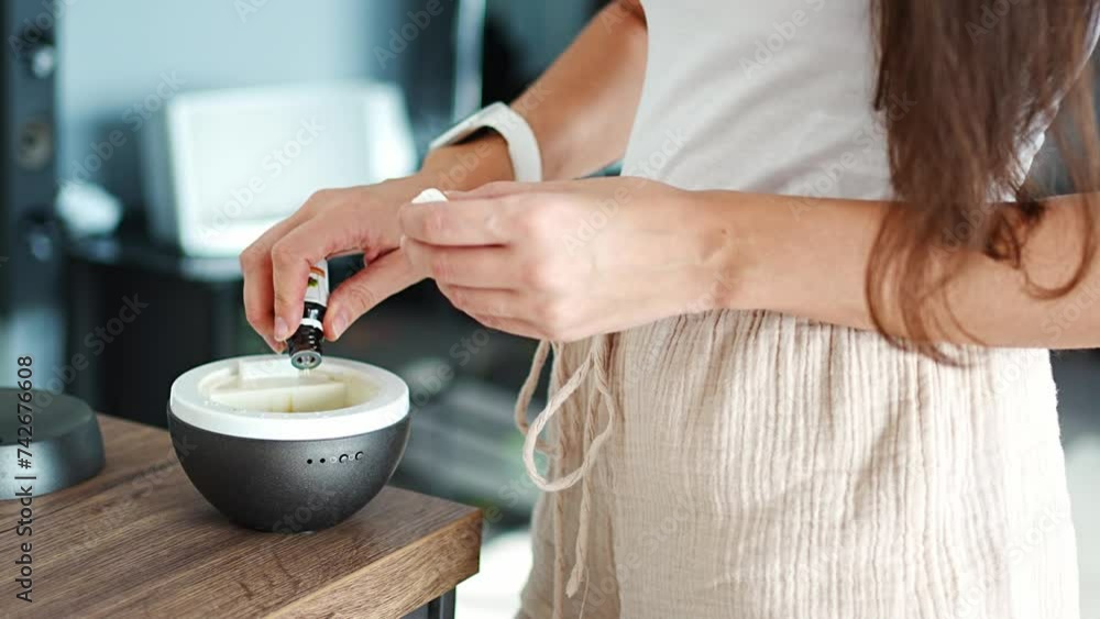 Woman adding oil to grey aroma oil diffuser on light table at home, turning on air freshener