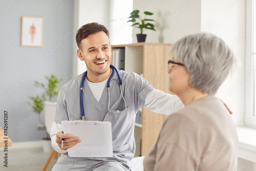 Obraz premium Young happy doctor, male nurse and old woman talking, supportive hand, taking social care. Man monitoring female resident of nursing home, patient in hospital, clinic, maintain elderly health, safety