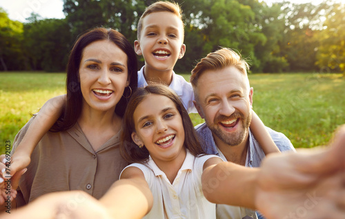 Selfie portrait of a happy smiling family of four in nature. Parents with kids spending time together and having fun in summer park. Little girl taking photo with mother  father and brother.