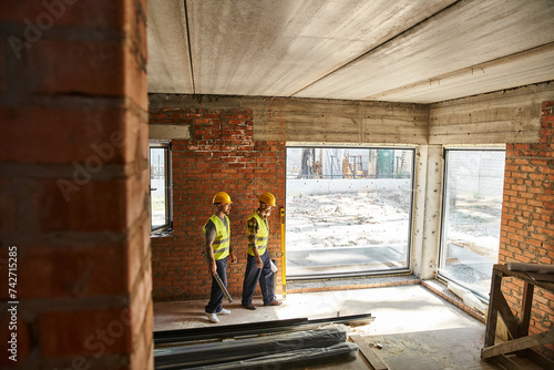 dedicated appealing men in safety vests working on site with blueprints and level, cottage builders