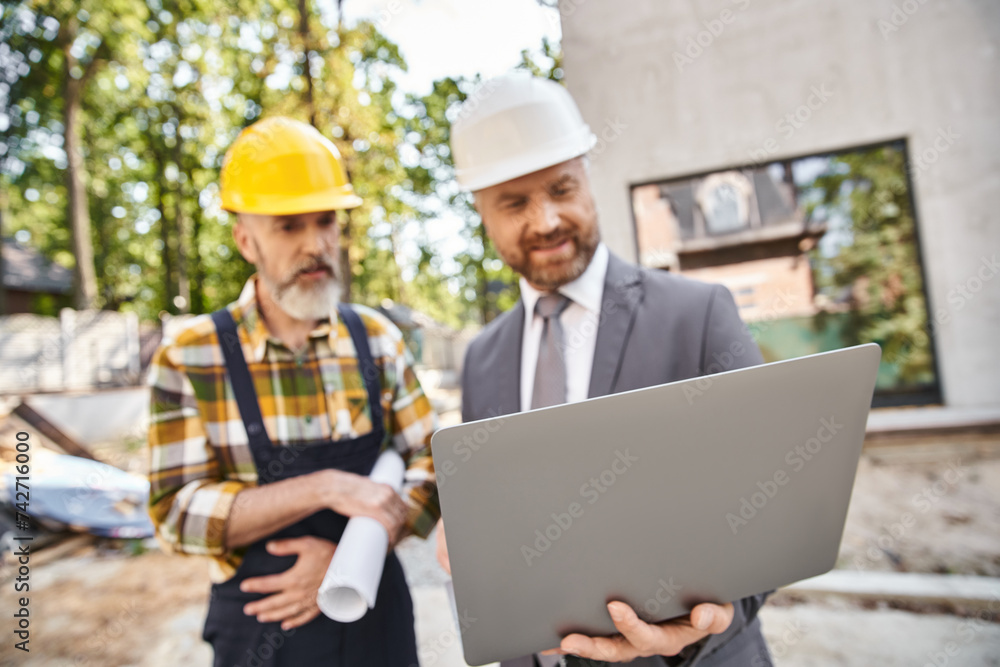 focus on laptop in hands of blurred architect and builder in safety helmets on construction site focus on laptop in hands of blurred architect and builder in safety helmets on construction site