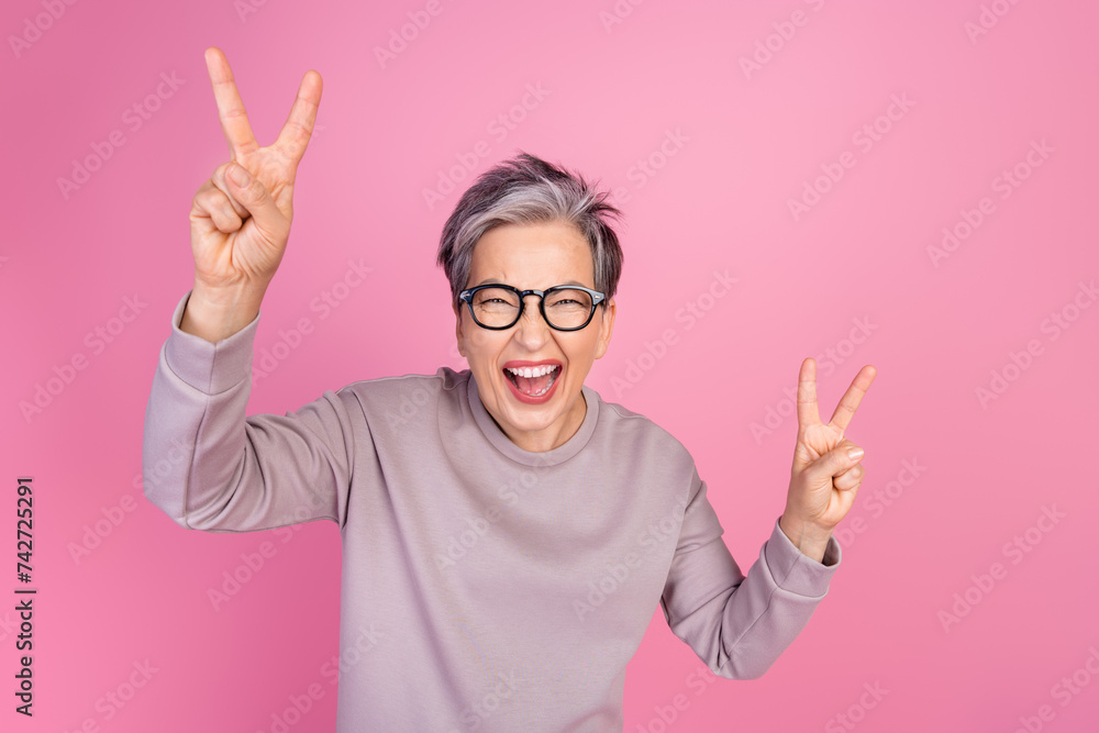 Photo of positive crazy funny senior woman wearing stylish clothes showing v-sign cool party isolated on pink color background Photo of positive crazy funny senior woman wearing stylish clothes showing v-sign cool party isolated on pink color background