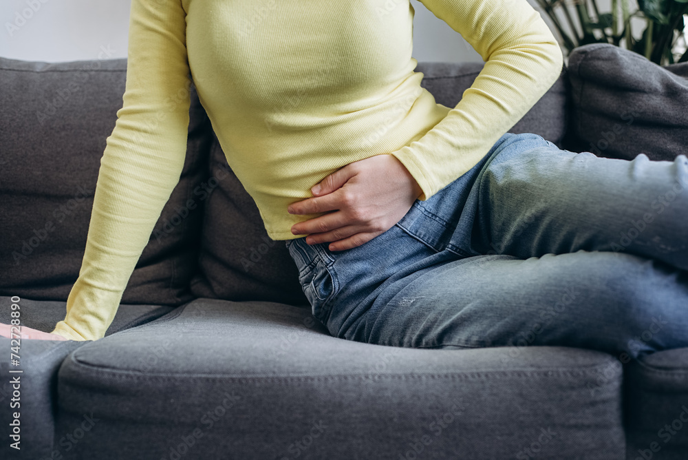 Ill female holding belly and feeling abdominal menstrual pain or bowel and digestion problems. Unhealthy young caucasian woman suffering from stomach ache sitting on couch. Food poisoning concept