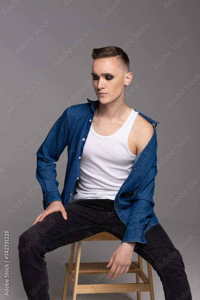 A young man in a blue shirt with rock musician makeup poses on a chair. A young man in a blue shirt with rock musician makeup poses on a chair.