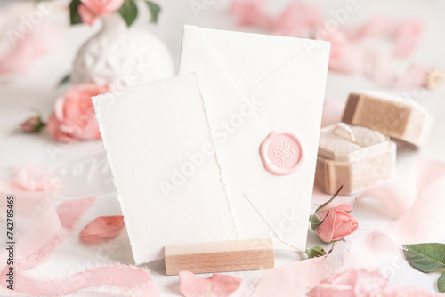 Card and sealed envelope near pink roses, petals and silk ribbons close up, wedding mockup