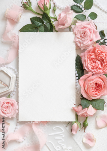 Vertical blank card near pink roses, engagement ring and silk ribbons top view, wedding mockup