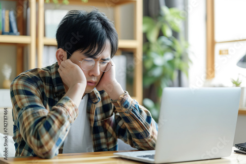 Exhausted Japanese man taking off eyeglasses and rubbing nose bridge, taking break in online work on laptop