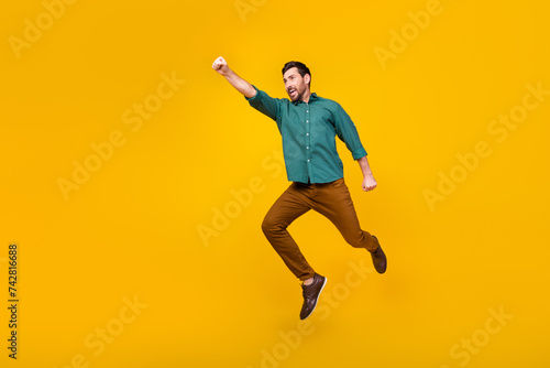 Full size photo of brave guy dressed dotted shirt brown trousers fly in empty space raising fist up isolated on yellow color background