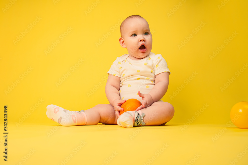 Beautiful baby girl holds orange sitting in studio on yellow background. Beautiful baby girl holds orange sitting in studio on yellow background.