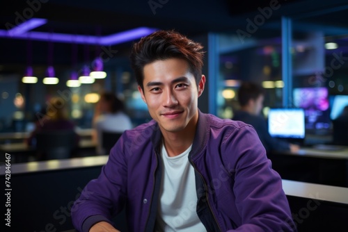 A young asian man with a pleasant smile, seated in a modern, dimly lit office with ambient blue lighting, which gives off a relaxed and contemporary vibe.