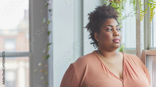 copy space, stockphoto, Plus size Black Woman standing in front of a window. Beautiful woman standing in front of a window in her home, watching outside. Big is beautiful. Body inclusive theme. Heavy 