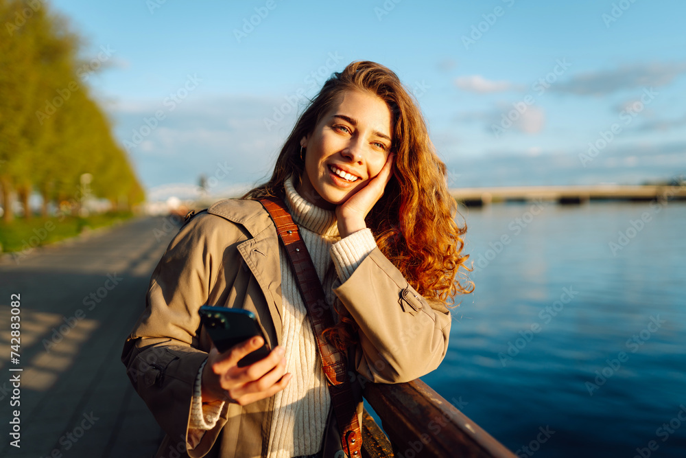 Young woman in coat enjoying views on embankment. Stylish woman with phone. Concept of vacation, technology, weekend.