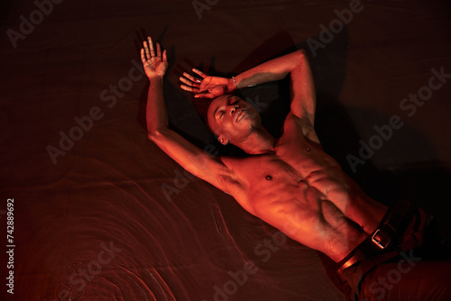 sexy african american man lying topless on water with closed eyes, dark lights, fashion concept