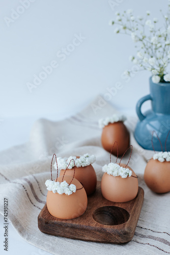 On a linen napkin there is a wooden egg cup with brown Easter eggs with flower wreaths and bunny ears. In the background there is a blue vase with gypsophila flowers. White background © Hanna