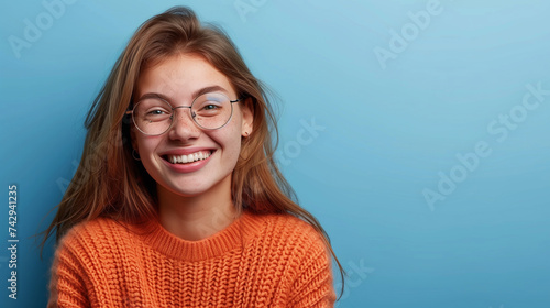 Happy pretty young woman wearing glasses looking at the camera with a big beaming friendly smile on blue with copy space .