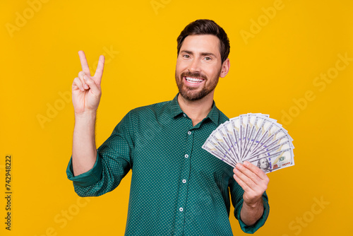 Photo of cheerful pleasant guy with beard dressed dotted shirt holding money showing v-sign symbol isolated on yellow color background photo