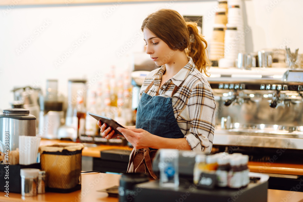 Smiling female barista takes an order from a tablet while standing at the bar counter in a coffee shop. Online order. Small business, people, takeaway and service concept. Smiling female barista takes an order from a tablet while standing at the bar counter in a coffee shop. Online order. Small business, people, takeaway and service concept.