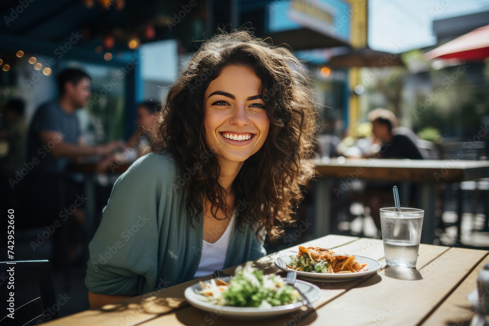 American young woman smiling while sitting outside at a table on a restaurant patio and eating delicious tacos.