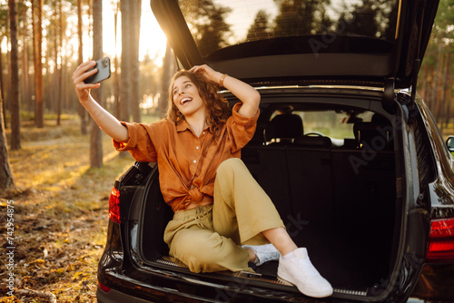 Сheerful woman traveler sitting in open trunk taking selfie on smartphone while traveling. Lifestyle, travel, tourism, nature, active life. © maxbelchenko