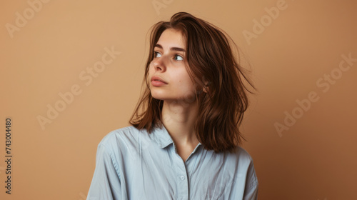 A young woman with shoulder-length hair gazes to the side against a neutral beige background, wearing a light blue shirt.
