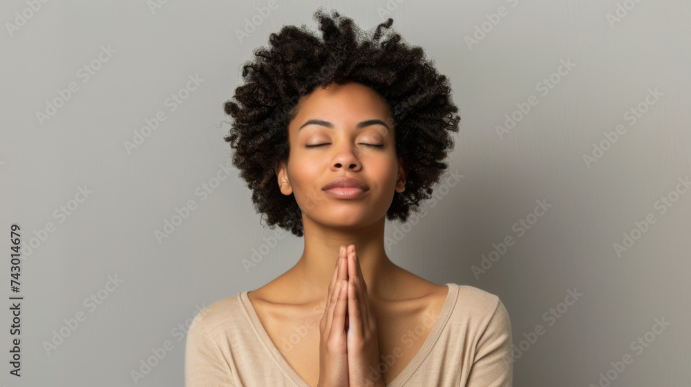 A serene individual with closed eyes is meditating, hands clasped together in a gesture of peace against a neutral background. - obrazy, fototapety, plakaty 