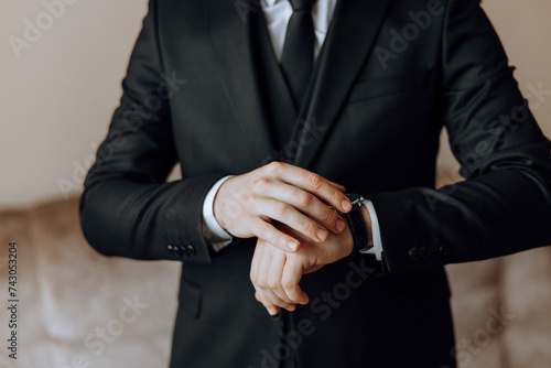 A close-up of a cropped frame of a man puts on a watch with a leather belt, is dressed in a stylish suit, a white shirt, wears a gold ring.