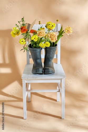 gardening, international women's day and floral design concept - flowers in rubber boots and straw hat on vintage chair over beige background