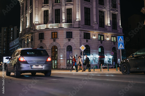 Group of well-dressed business people crossing a busy street in the evening with cars and classical architecture in the background.